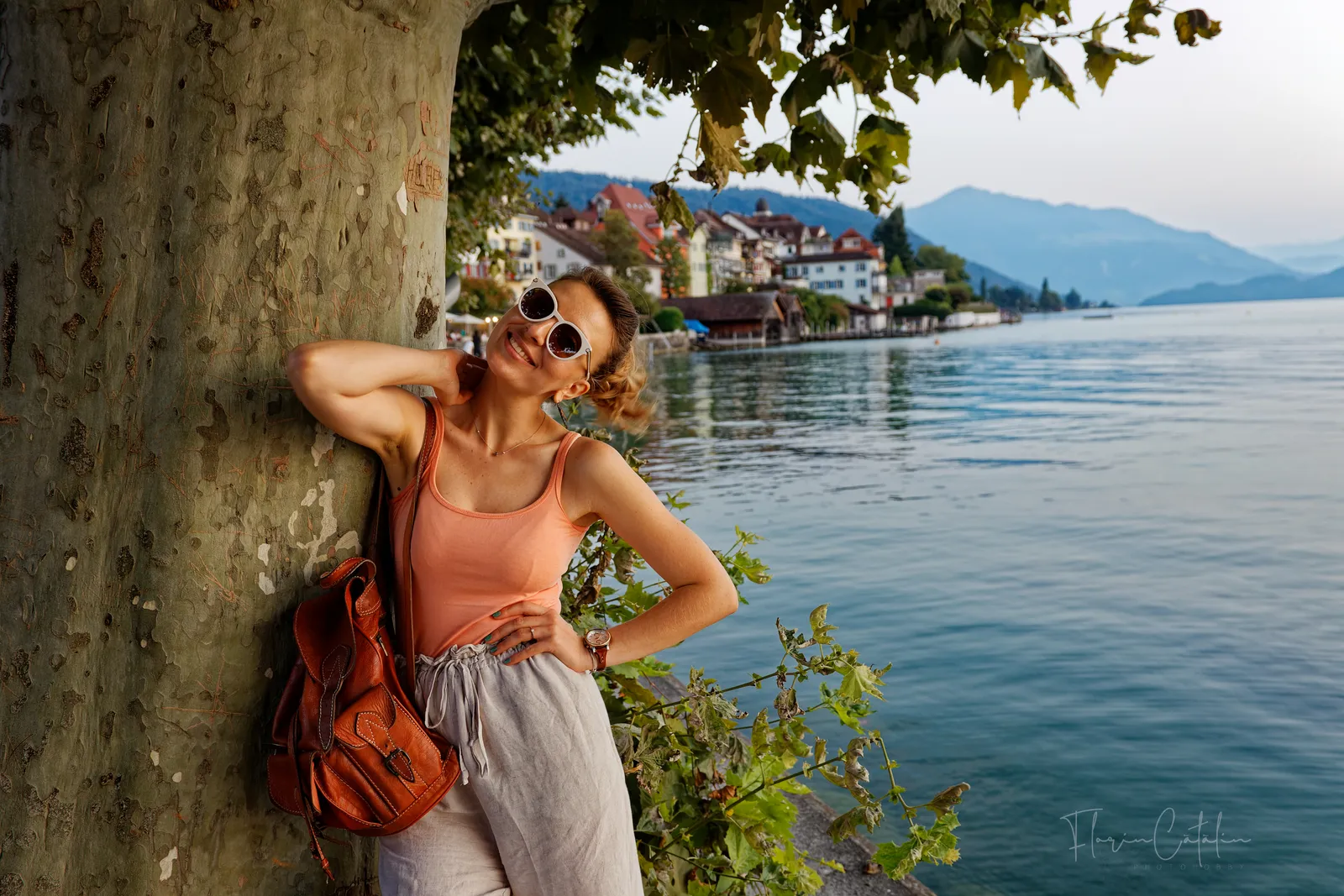 Reise-Fotoshooting am Schweizer Seeufer – Frau lehnt lächelnd an einem Baum, im Hintergrund Dorf und Berge