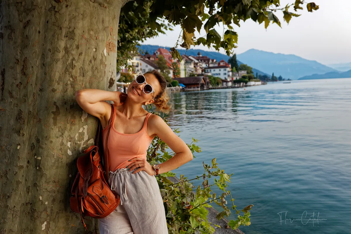 Reise-Fotoshooting am Schweizer Seeufer – Frau lehnt lächelnd an einem Baum, im Hintergrund Dorf und Berge