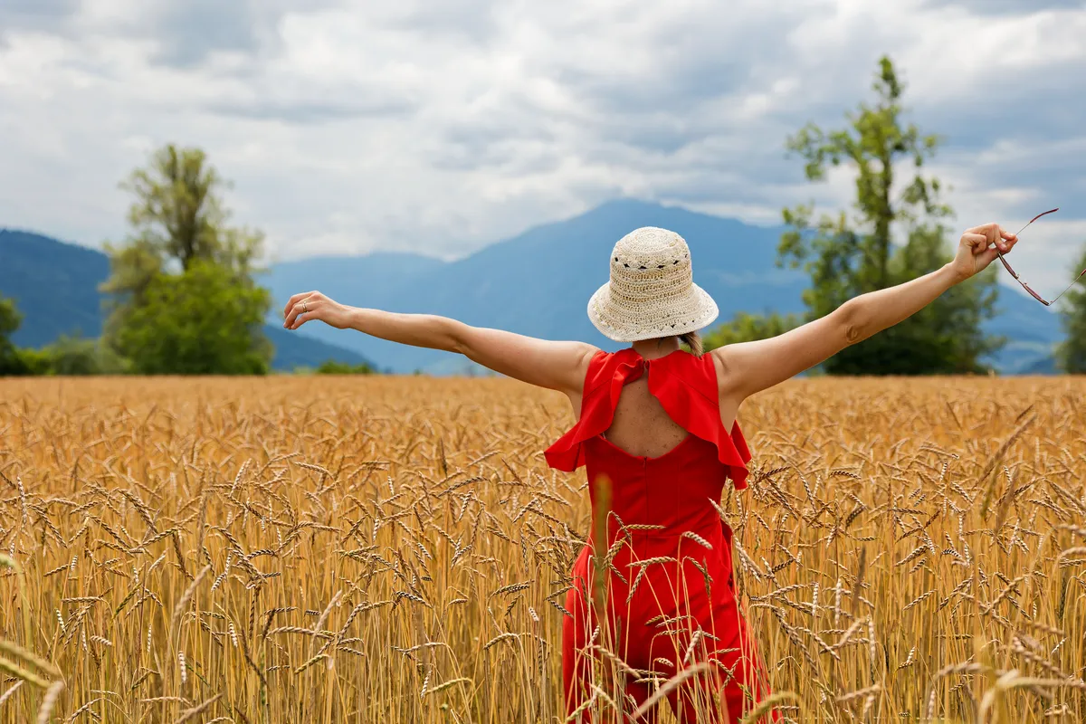 Reise-Fotoshooting im Weizenfeld – Frau im roten Kleid streckt die Arme aus, Berge im Hintergrund